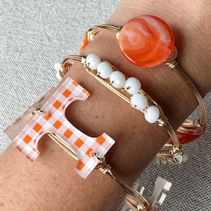 Op de afbeelding: Gouden draad armbanden met oranje en witte geruite, oranje swirl en witte kralen bedels. De armbanden zijn gestapeld op de pols van een persoon.