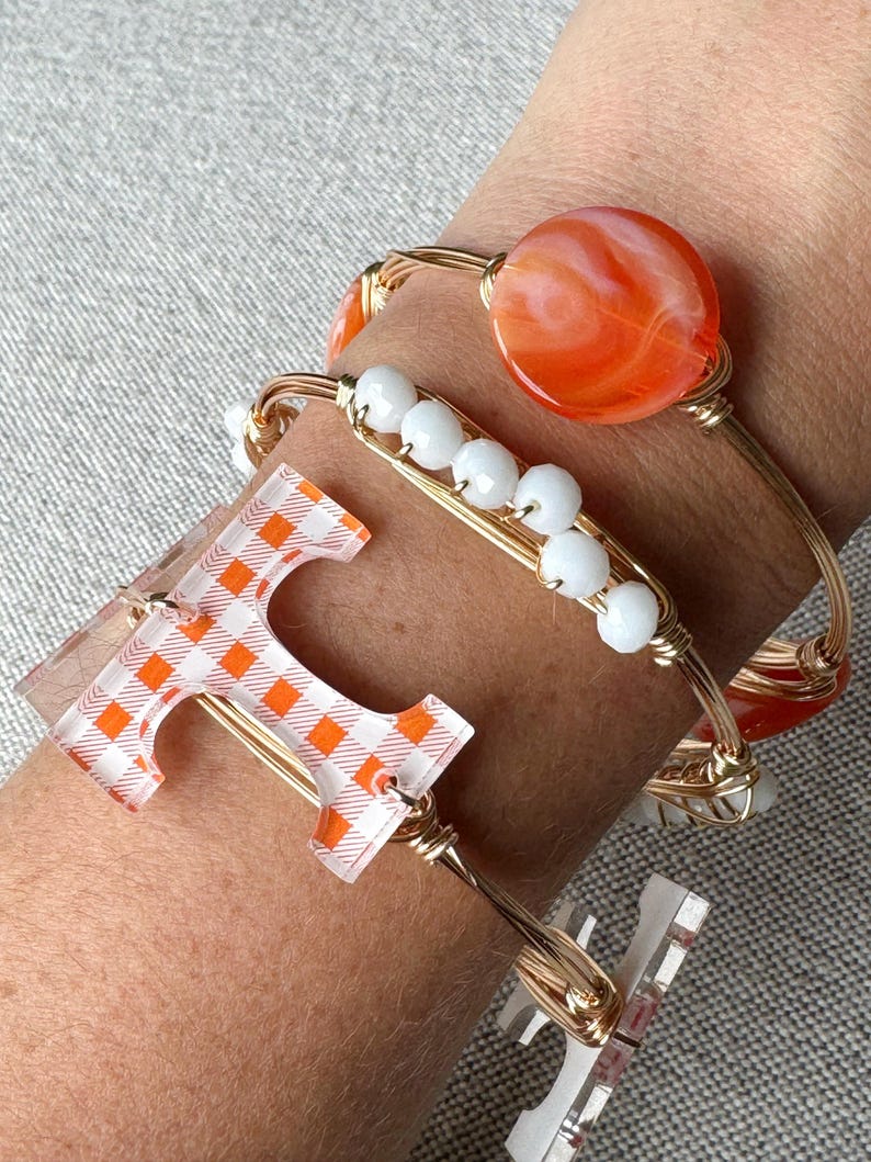 Op de afbeelding: Gouden draad armbanden met oranje en witte geruite, oranje swirl en witte kralen bedels. De armbanden zijn gestapeld op de pols van een persoon.
