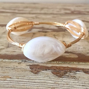 May include: A gold-toned wire bracelet featuring three large, oval-shaped white beads. The beads have a marbled appearance and are securely wrapped with the gold wire. The bracelet is displayed on a light brown, textured surface.