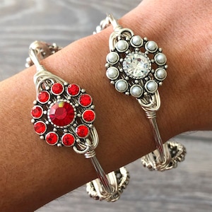 Könnte beinhalten: Zwei silberne, drahtgewickelte Armbänder mit einem roten Strassstein und einem weißen Perlenmuster. Die Armbänder befinden sich am Handgelenk einer Person.