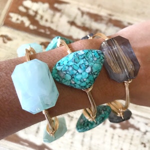 Puede incluir: Cuatro pulseras de alambre dorado con piedras de diferentes colores. Las piedras son turquesa, azul claro y gris. Las pulseras est&aacute;n apiladas en la mu&ntilde;eca de una persona.