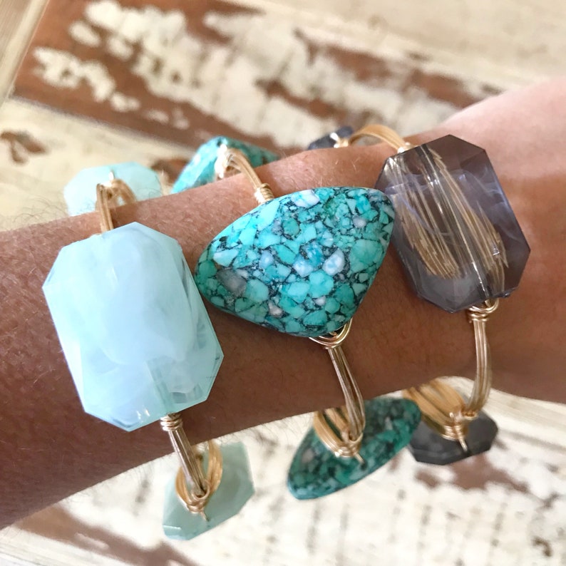 Puede incluir: Cuatro pulseras de alambre dorado con piedras de diferentes colores. Las piedras son turquesa, azul claro y gris. Las pulseras est&aacute;n apiladas en la mu&ntilde;eca de una persona.