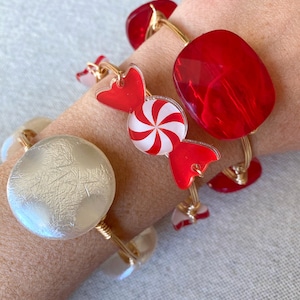 May include: Three gold bangle bracelets with red and white candy cane and red and white peppermint designs. One bracelet has a white round charm.