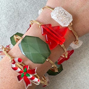May include: Three gold wire bracelets with Christmas-themed charms. One bracelet features a candy cane with a red bow, another has a red Christmas tree, and the third has a green gemstone. The bracelets are adorned with clear crystal accents.