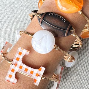 Op de afbeelding: Een set van vier gouden draad armbanden met verschillende bedels. De bedels omvatten een witte marmerachtige bedel, een bruine voetbalbedel, een oranje glazen bedel en een transparante bedel met een geruit oranje en wit patroon. De bedel met het geruite patroon is in de vorm van de letter "T".