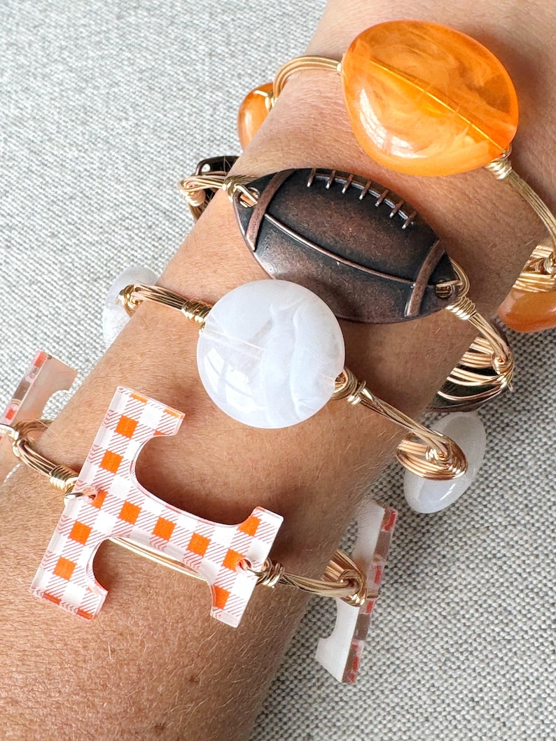 Op de afbeelding: Een set van vier gouden draad armbanden met verschillende bedels. De bedels omvatten een witte marmerachtige bedel, een bruine voetbalbedel, een oranje glazen bedel en een transparante bedel met een geruit oranje en wit patroon. De bedel met het geruite patroon is in de vorm van de letter "T".