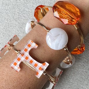 Op de afbeelding: Een set van vier gouden draad armbanden met verschillende bedels. Een armband heeft een witte marmerachtige bedel, een andere heeft een grote oranje glazen bedel, en de derde heeft een heldere bedel met een geruit oranje en wit patroon. De vierde armband heeft een heldere bedel met de letter "T" in een geruit oranje en wit patroon.