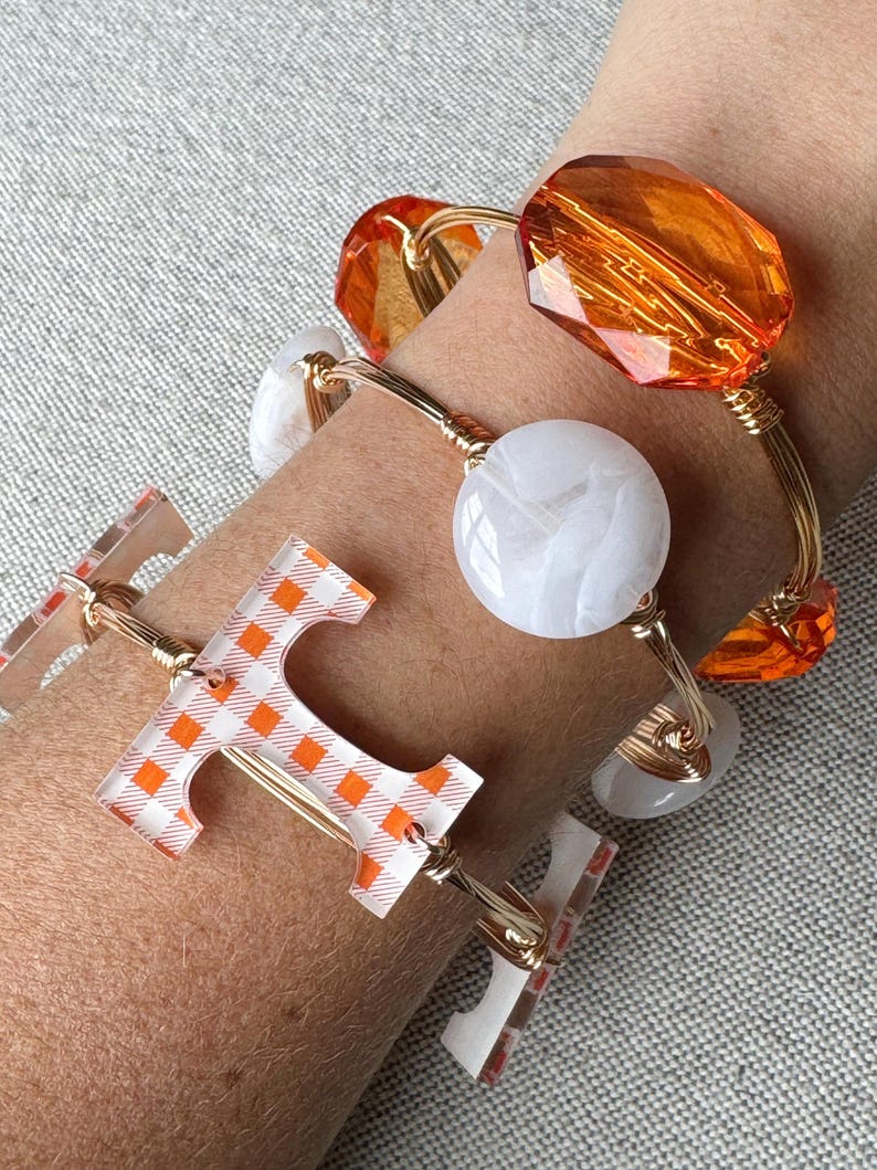 Op de afbeelding: Een set van vier gouden draad armbanden met verschillende bedels. Een armband heeft een witte marmerachtige bedel, een andere heeft een grote oranje glazen bedel, en de derde heeft een heldere bedel met een geruit oranje en wit patroon. De vierde armband heeft een heldere bedel met de letter "T" in een geruit oranje en wit patroon.