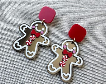 May include: A pair of dangle earrings featuring gingerbread man charms with a brown finish, white icing details, red bow ties, and two peppermint candy buttons. Each charm hangs from a solid red square stud top with a gold-tone metal jump ring.