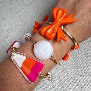 Può includere: Tre braccialetti colorati. Uno presenta un fiocco arancione, un altro ha un globo bianco e il terzo un design geometrico rosa, arancione e bianco. Tutti sono su filo d'oro.