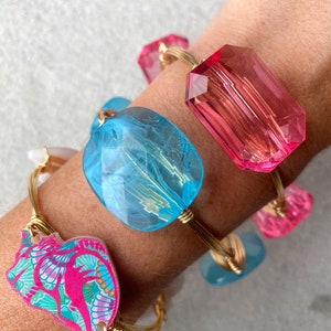 May include: A set of four gold wire bracelets with colorful charms. The charms include a pink and blue butterfly, a light blue oval, a pink rectangle, and a light blue rectangle.