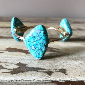 Puede incluir: Un brazalete de alambre dorado envuelto con tres piedras turquesa. Las piedras son de color azul y verde moteado.