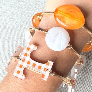 Op de afbeelding: Een set van drie gouden draad armbanden met verschillende bedels. De bedels omvatten een witte marmerachtige bedel, een grote oranje bedel en een transparante bedel met een geruit patroon.