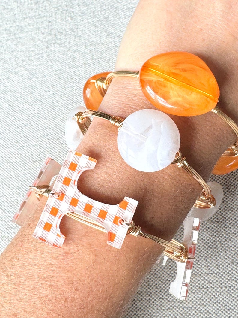 Op de afbeelding: Een set van drie gouden draad armbanden met verschillende bedels. De bedels omvatten een witte marmerachtige bedel, een grote oranje bedel en een transparante bedel met een geruit patroon.