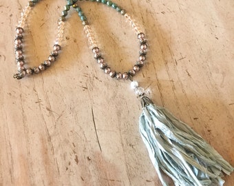 Op de afbeelding: Een lange ketting met een kleurverloop in turquoise, crème en roze. De ketting heeft een kwast hanger gemaakt van lichtblauwe stof.