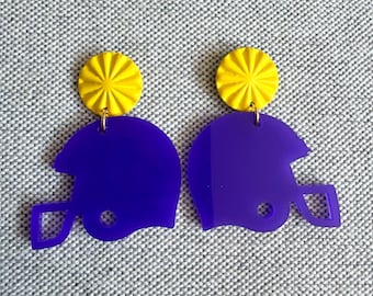 May include: A pair of earrings featuring a yellow circular top with a starburst design, connected to a purple football helmet shape. The earrings are displayed on a gray textured surface.