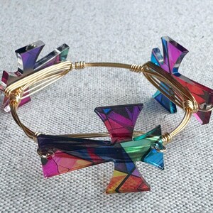 May include: A gold-toned wire bangle bracelet featuring four cross-shaped charms. The charms are made of iridescent, multi-colored material, displaying hues of pink, purple, blue, and orange. The bracelet is set against a neutral, textured background.