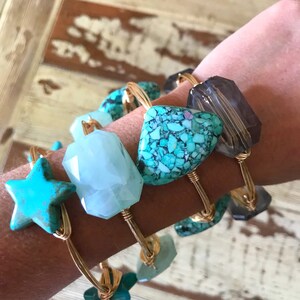 Puede incluir: Un conjunto de cuatro pulseras de alambre dorado con acentos de piedras preciosas turquesa y transparentes. Las pulseras presentan una estrella, una piedra rectangular, una piedra en forma de diamante y una piedra redonda.