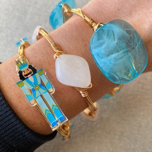 Op de afbeelding: Drie gouden draadgewikkelde armbanden met blauwe en witte harscharms. Een bedel heeft een notenkrakerontwerp, een andere is een witte ovaal en de derde is een blauwe swirl.