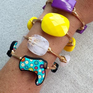May include: A collection of gold wire bracelets adorned with colorful beads and charms. The bracelets feature a variety of shapes, including a purple rectangle, a yellow faceted bead, a clear faceted bead, and a turquoise boot charm with a floral pattern.