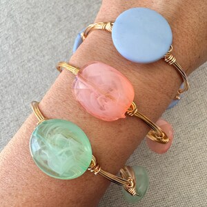 Op de afbeelding: Drie goudkleurige draadarmbanden met kleurrijke, doorschijnende kralen. Eén armband heeft een lichtblauwe schijfvormige kraal, een andere heeft een roze ovale kraal en de derde heeft een groene ovale kraal. De armbanden zijn op een pols gestapeld.