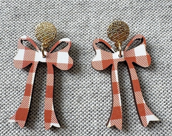 May include: A pair of bow-shaped earrings. The earrings feature a red and white gingham pattern on a wooden base. The top of each earring has a small, textured, gold-colored disc.