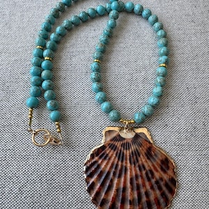 May include: A turquoise beaded necklace featuring a large, brown and tan seashell pendant. The necklace has gold-coloured accents and a gold clasp. The seashell pendant has a detailed, fan-like pattern.