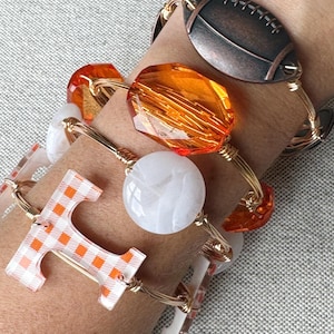 Op de afbeelding: Vier gouden draad armbanden met verschillende bedels. De bedels omvatten een voetbal, een grote oranje facetgeslepen kraal, een witte ronde kraal en een letter "T" met een wit en oranje geruit patroon.