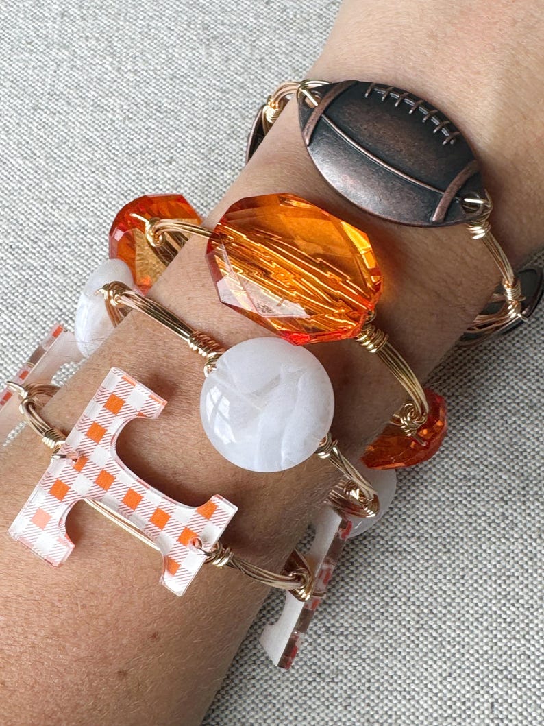 Op de afbeelding: Vier gouden draad armbanden met verschillende bedels. De bedels omvatten een voetbal, een grote oranje facetgeslepen kraal, een witte ronde kraal en een letter "T" met een wit en oranje geruit patroon.