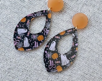 May include: A pair of Halloween-themed earrings. The earrings feature a teardrop shape with a black background and a pattern of ghosts, pumpkins, candy, and spiderwebs. The top of each earring is a small, round, orange stud.