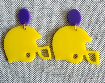 May include: A pair of yellow football helmet earrings with purple studs. The earrings are made of a shiny material and have a small hole on the side of the helmet. The earrings are on a gray background.
