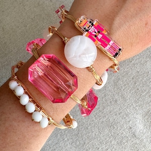 May include: A set of gold wire bracelets with pink and white glass beads and charms. The charms include a pink nutcracker, a white round bead, and a pink octagon-shaped bead.