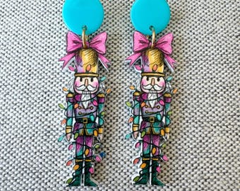 May include: A pair of dangle earrings featuring a nutcracker illustration wrapped in colorful string lights. Each earring has a solid turquoise circular stud at the top, connected to a nutcracker figure wearing a pink bow, purple uniform, and green boots.