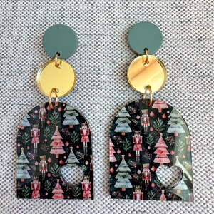 Puede incluir: Un par de pendientes festivos. Cada pendiente presenta un círculo verde azulado, un disco dorado y un arco negro con un patrón de árbol de Navidad y cascanueces. Una forma de corazón está recortada en la parte inferior de cada arco.