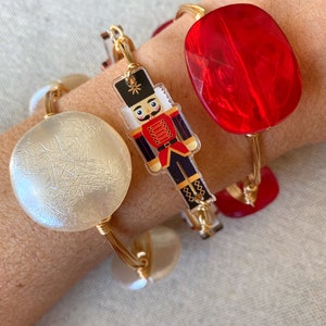 May include: Three gold wire bracelets with different charms. One bracelet has a white round charm, another has a red round charm, and the third has a clear charm with a cartoon nutcracker.