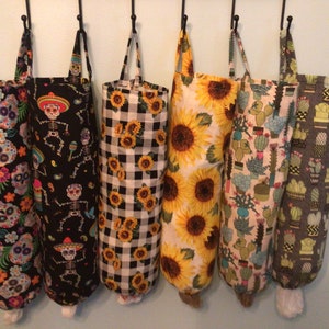 May include: Six fabric grocery bag holders with various patterns, including sugar skulls, skeletons, sunflowers, and cacti. Each holder is cylindrical with a loop handle and a white bottom. The holders hang from black hooks.