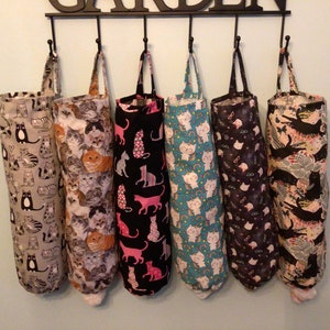 May include: Six fabric bag dispensers with various cat-themed patterns hang from a black metal rack. The patterns include cats in gray, orange, and black, with some bags featuring pink cats on black. Each bag has a loop for hanging.
