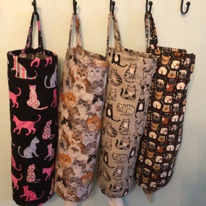 May include: Four fabric bag dispensers hanging on black hooks. The bags feature various patterns, including cats in pink, white, and blue on black, a collage of cat faces, cats on a gray background, and dog faces on a dark background.