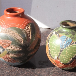 May include: Two round ceramic vases with narrow openings. The larger vase features a terracotta top and dark green body with carved leaf patterns. The smaller vase displays a prominent green and blue butterfly design against a dark green background.