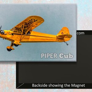 Könnte beinhalten: Ein gelbes Piper Cub Flugzeug mit schwarzen Markierungen und der Nummer "2989C" an der Seite. Das Flugzeug befindet sich im Flug vor einem hellblauen Himmel. Die Worte "PIPER Cub" sind unter dem Flugzeug gedruckt.