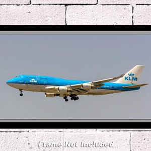 Op de afbeelding: Een ingelijste print van een KLM Boeing 747 vliegtuig in vlucht tegen een heldere hemel. Het vliegtuig is voornamelijk blauw en wit, met het KLM-logo op de staart. De lijst is zwart, de achtergrond een witte bakstenen muur.