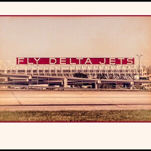 May include: A vintage-style photo of a red sign with the words "FLY DELTA JETS" against a light brown background. The sign is mounted on a metal structure with a series of white beams.
