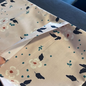 May include: A light pink fabric pouch featuring a repeating pattern of dark navy blue leaves, small teal dots, and stylized white flowers with orange centers. The pouch has a folded top edge and a smooth, lightweight textile construction.