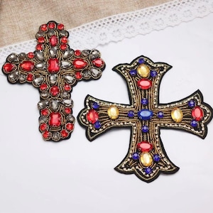 May include: Two decorative cross patches. One cross is adorned with red and gray gemstones, the other with red, blue, and yellow gemstones. Both crosses have intricate beadwork and are set against a neutral background.