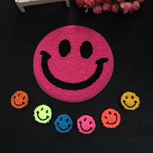 Peut inclure: Un ensemble de patchs smiley colorés et un grand tapis smiley rose. Les patchs sont de différentes couleurs, notamment orange, vert, bleu, rose et jaune. Le tapis a un sourire et des yeux noirs.