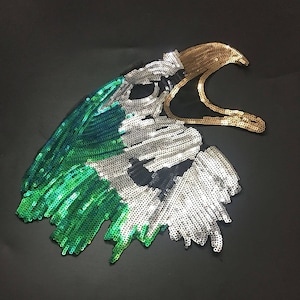 May include: A green, white and gold sequined eagle head applique. The eagle's beak is open and the eyes are black.