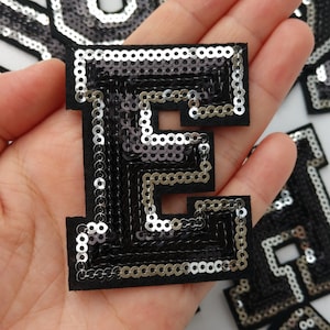 Może przedstawiać: Naszywka z literą E wykonana z srebrnych i czarnych cekinów. Naszywka jest wykonana z cekinów przyszytych do czarnego filcowego podkładu.