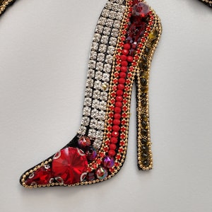 Puede incluir: Un zapato de tacón alto rojo y plateado hecho de pedrería y cuentas. El zapato es una pieza decorativa y no está diseñado para ser usado.
