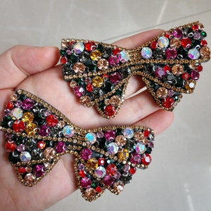 May include: Two decorative bow-shaped embellishments. Each bow is adorned with an array of colourful rhinestones in shades of red, green, yellow, and iridescent hues. The bows have a gold-coloured border.