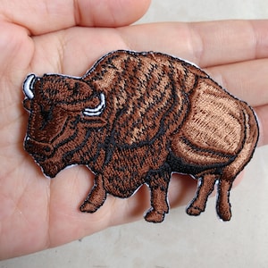 以下が含まれることがあります： 詳細なステッチが施された茶色のバイソンの刺繍パッチ。パッチには白い縁があり、生地にアイロンをかけたり縫い付けたりするように設計されています。バイソンは、質感と色に焦点を当てた、リアルなスタイルで描かれています。